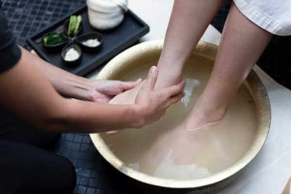 Mujer recibiendo baño de pies con hierbas y sales especiales en un spa. Sus pies están sumergidos en un cuenco grande y están siendo masajeados por una terapeuta.