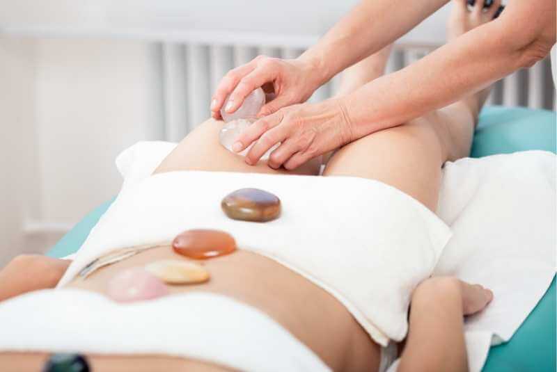 Terapeuta realizando amasamientos y fricciones con piedras de cuarzo para reducir la celulitis en el musla de una clienta.