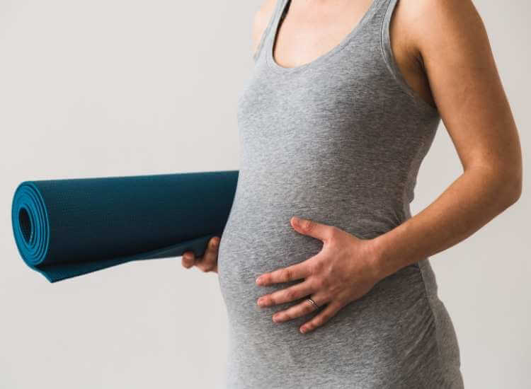 Mujer embarazada con camiseta atlètica cargando una colchoneta de yoga.